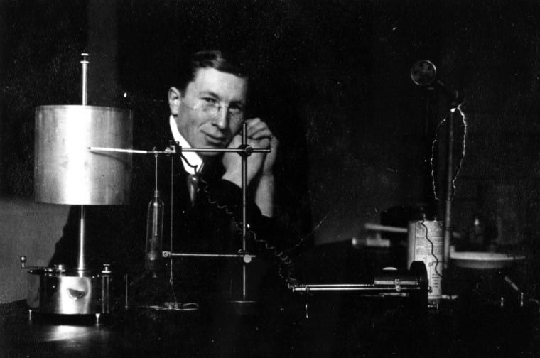 Black and white photograph showing man sitting at a lab table, behind lab equipment, and looking toward the camera.