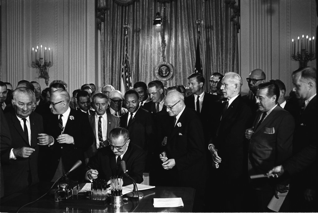 President Lyndon B. Johnson signs the 1964 Civil Rights Act as Martin Luther King, Jr. and others look on.East Room, White House, Washington DC, on 7 February 1964.