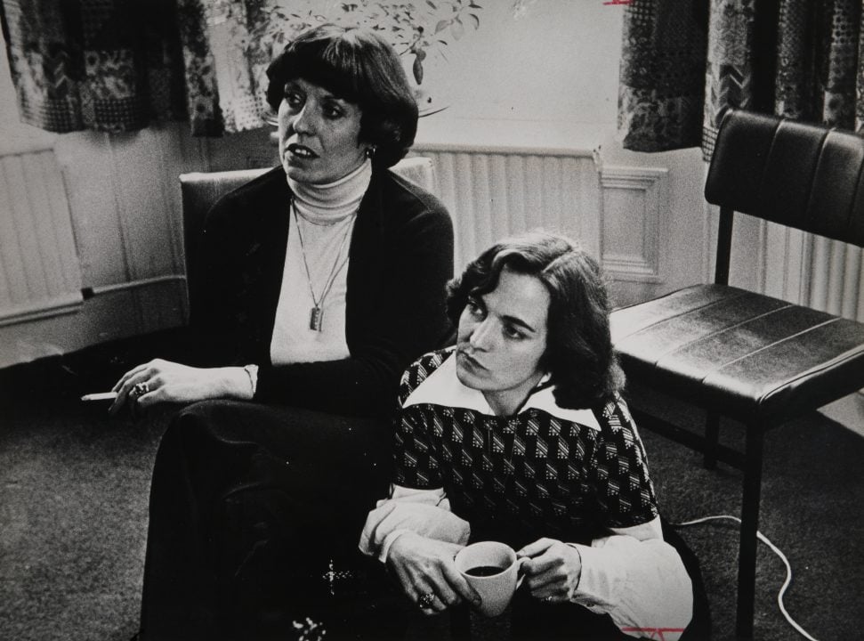 Two women sitting in a room, one is smoking, the aother drinking coffee